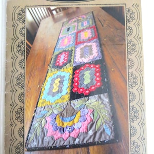 May include: A colorful, handmade table runner with a patchwork design. The runner features squares of various colors and patterns, including red, blue, green, and pink. The text "Max & Louise Pattern Co. Small Treasures" is visible at the top.