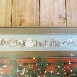 May include: A framed cross-stitch sampler featuring the alphabet, snowmen, and winter-themed elements. The letters are stitched in a brown thread against a light blue background. The frame is a muted brown color. Decorative greenery is visible at the bottom.