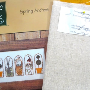 May include: A cross stitch pattern for a Spring Arches design. The pattern includes five arched windows with different scenes, including a bunny, a bird, and a basket of flowers. The pattern is printed on a white background with a brown wooden frame. The pattern is accompanied by a piece of beige linen fabric.