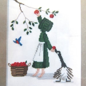 May include: Cross-stitch embroidery of a girl in a green dress and white apron picking apples from a tree. A goose walks beside her, and a basket of apples sits on the ground.