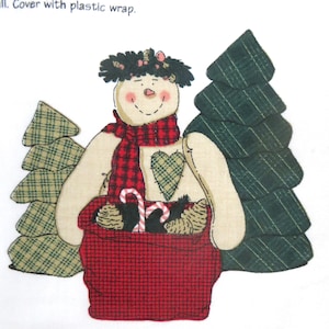 May include: A fabric snowman with a red plaid scarf and a red basket filled with candy canes and pinecones. The snowman has a green heart-shaped patch and is flanked by green plaid Christmas trees. The image has a festive, handmade feel.