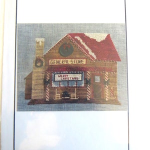 May include: Cross stitch pattern for a Christmas village general store. The store is brown with a red roof and a sign that says "General Store". The window has a sign that says "Merry Christmas".