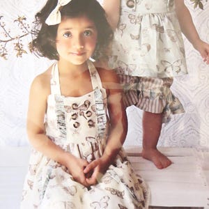 May include: Two young girls wearing vintage-style dresses with butterfly patterns. The dresses are cream-colored with brown butterfly prints and ruffled details. One girl is seated, wearing a halter dress, while the other stands in a matching top and striped shorts. The text "Ginger vintage chic top & dress" is visible.