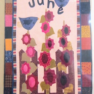 May include: A fabric wall hanging with the word "June" and two blue birds above a row of sunflowers. The sunflowers are pink and burgundy, with green leaves, and sit behind a brown picket fence. The border is made of various fabric squares.