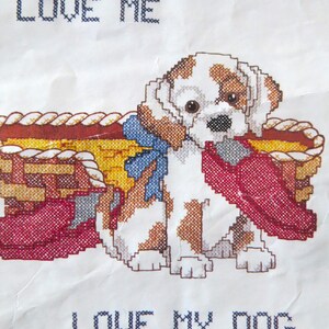 May include: Cross-stitch pattern featuring a puppy with brown and white spots, holding a red item, and sitting next to a basket. The text "LOVE ME" and "LOVE MY DOG" are stitched above and below the image.