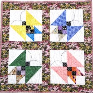 May include: A handmade quilt featuring four square blocks with butterfly designs. Each block has a white background with colorful triangles and patterned squares. The quilt is bordered with a floral fabric and a pink edge.