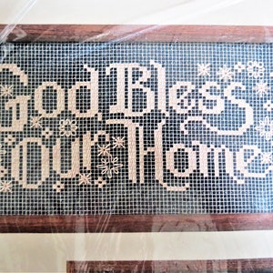 May include: A framed cross-stitch with the text "God Bless Our Home" in a beige thread on a black mesh background. The frame is made of dark wood.