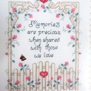 May include: Cross-stitch artwork featuring the quote "Memories are precious when shared with those we love." The design includes a white picket fence, pink roses, a butterfly, and a decorative border with a heart.