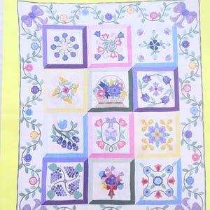 Puede incluir: Un tapiz de pared acolchado con el texto "GARDEN WINDOW" en la parte superior. El edredón presenta nueve bloques cuadrados con diseños florales en varios colores, rodeados por un borde de vides, lazos y flores. El nombre y el año del artista están en la parte inferior.