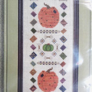 May include: A framed cross-stitch featuring two orange pumpkins and a green pumpkin, surrounded by colorful geometric patterns. The design includes floral and diamond shapes, perfect for fall decor.