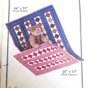Puede incluir: Un patrón para colchas de muñecas, con un oso de peluche sobre dos colchas. La colcha más grande mide 40,6 cm x 53,3 cm con un borde azul y un patrón de diamantes rojos y blancos. La colcha más pequeña mide 25,4 cm x 38,1 cm.