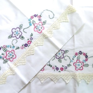 May include: Two white pillowcases with embroidered floral designs and a crocheted lace trim. The flowers are pink, blue, and purple.
