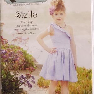 May include: A sewing pattern package for a child's one-shoulder dress named "Stella." The package features a young girl wearing a light purple dress with a ruffled neckline. The package includes the text "Violette Field Threads."
