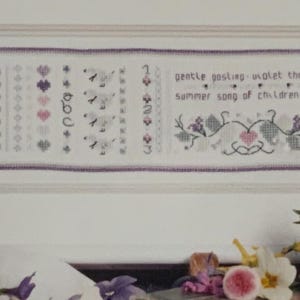 May include: Framed cross-stitch sampler with a white background and a purple border. The sampler features various patterns, including hearts, flowers, and birds, along with the text "gentle posting-violet thread summer song of children led."