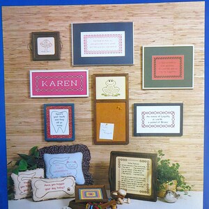May include: A collection of framed cross-stitch embroidery designs with various themes, including animals, friendship, and inspirational quotes. The designs are displayed on a wood-panelled wall with a corkboard and a tray of embroidery supplies.