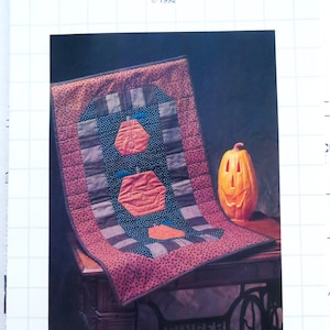 May include: A quilt pattern featuring a patchwork design of pumpkins. The quilt is displayed on a table with a carved pumpkin beside it. The pattern is titled "Pumpkins Patched" and is a "Master Piece" by Jill Reber, copyright 1992.