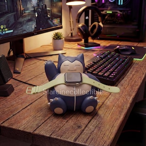 May include: A blue and white Snorlax-shaped Apple Watch charging stand with a light green watch strap. The stand is on a wooden desk with a keyboard, mouse, and computer monitor in the background. The text "Starcinecollectible" is visible.