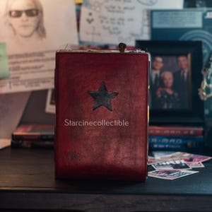 Può includere: Un diario in pelle rossa con una stella nera. Il diario presenta il testo "Starcinecollectible". È appoggiato su una superficie scura, con una foto incorniciata e altri oggetti sullo sfondo.