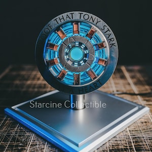 May include: A silver-colored collectible featuring a circular design with blue and orange accents, reminiscent of a reactor. The text "PROOF THAT TONY STARK HAS A HEART" is inscribed around the edge. It is mounted on a square base with the text "Starcine Collectible".