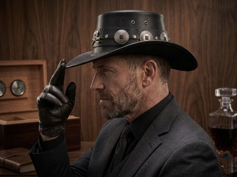 May include: Black leather cowboy hat with silver embellishments, including skull and crossbones and circular designs. The hat has a wide brim and is worn by a person wearing a black suit and gloves. The hat is a western-themed accessory.