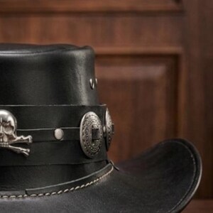 May include: Black leather cowboy hat with silver skull and crossbones emblem and decorative silver studs. The hat features a wide brim and a stitched brim edge. A stylish accessory.