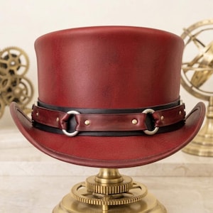 May include: A burgundy top hat with a black band and silver-colored metal accents. The hat is displayed on a brass stand with a gear-like base. The hat has a vintage, steampunk aesthetic.