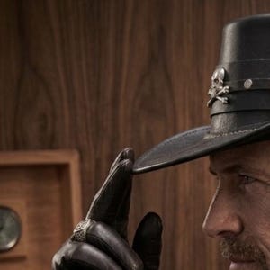May include: Black leather hat with a flat brim, adorned with a silver skull and crossbones emblem and a decorative circular button. The hat is part of a western-themed collection.