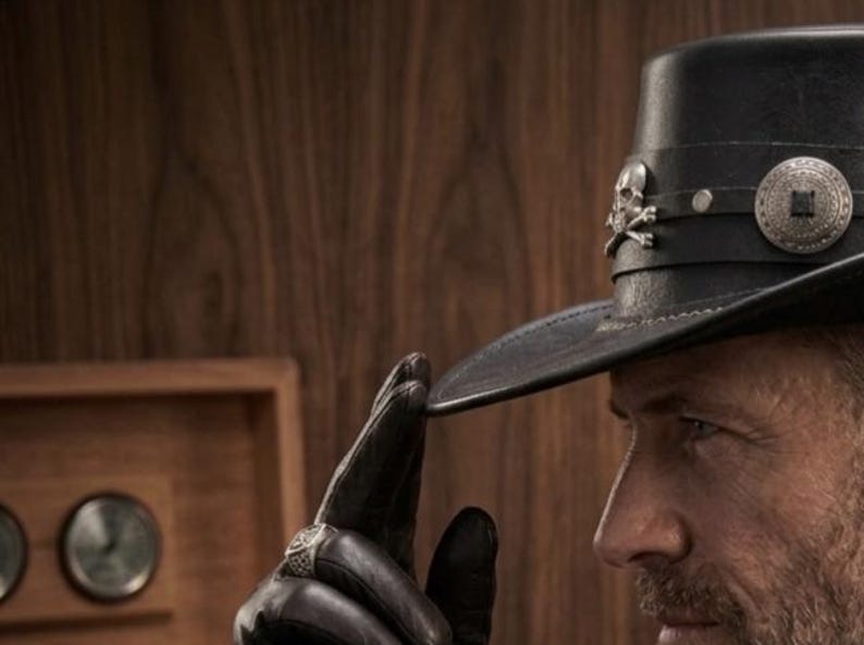 May include: Black leather hat with a flat brim, adorned with a silver skull and crossbones emblem and a decorative circular button. The hat is part of a western-themed collection.