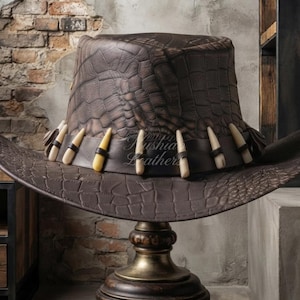 May include: A dark brown leather hat with a textured crocodile skin pattern. The hat features a wide brim and a dark brown leather band adorned with a series of light-colored, tooth-shaped decorative elements spaced evenly around the crown.