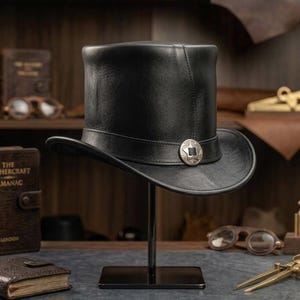Op de afbeelding: Een zwarte leren hoge hoed met een zilverkleurig embleem op de band. De hoed wordt op een standaard getoond, met boeken en vintage gereedschap op de achtergrond. De rand van de hoed heeft een diameter van ongeveer 30 cm.
