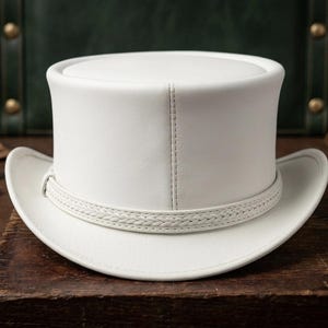 May include: A white top hat with a braided band around the crown. The hat is made of a smooth material and sits on a wooden surface. The hat has a classic design.