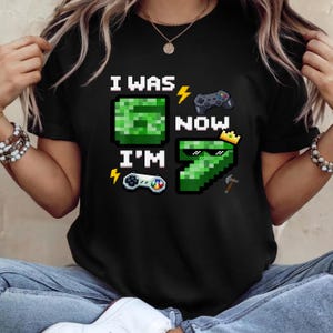 Könnte beinhalten: Schwarzes T-Shirt mit einem pixeligen Design. Das Design enthält den Text "I WAS 6 NOW I'M 7" mit pixeligen Bildern eines Creepers, eines Controllers, einer Krone und einer Spitzhacke. Das T-Shirt wird von einer Person getragen.