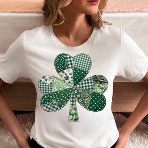 May include: White t-shirt with a large shamrock design. The shamrock is made up of different green patterns, such as polka dots, gingham, and floral prints, giving a patchwork look. The shirt has short sleeves and a crew neck.