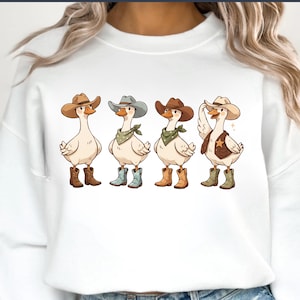 May include: White sweatshirt with a design of four cartoon geese in cowboy attire. Each goose has a cowboy hat, boots, and a bandana or vest. The geese are in shades of brown, blue, and green. The illustration has a fun, lighthearted feel.