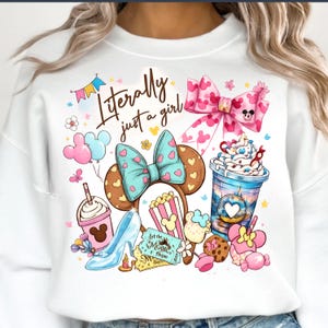 Pode incluir: Camisola branca com um gráfico colorido com um laço da Minnie, orelhas do Mickey, um sapato da Cinderela, pipocas e uma chávena de café. O texto "Literally just a girl" também está incluído.