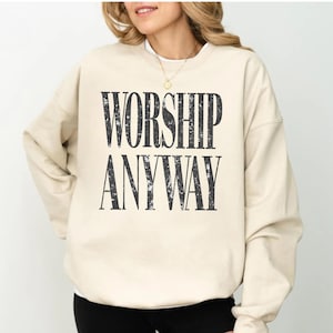 May include: A beige sweatshirt with the words "WORSHIP ANYWAY" printed in a distressed black font. The sweatshirt has a crew neck and long sleeves, offering a casual and comfortable style.