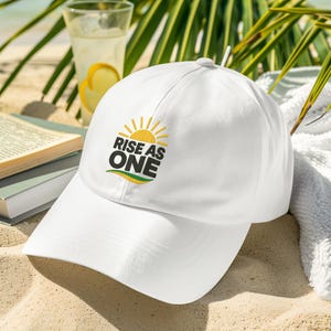Może przedstawiać: Biała czapka bejsbolowa z napisem "RISE AS ONE" i grafiką słońca. Czapka leży na piaszczystej plaży ze szklanką lemoniady, książką i liśćmi palmowymi w tle.