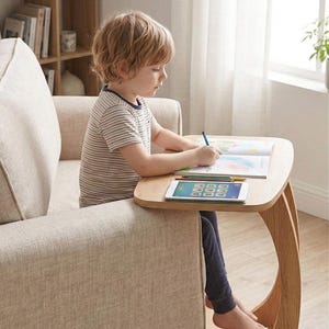 May include: A light wood side table with a curved leg design. The table is positioned next to a beige sofa. A child is seated on the sofa, drawing on a coloring book and using a tablet. The room has a neutral color palette.