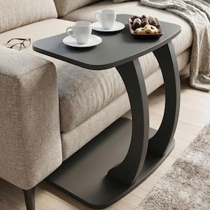 May include: A dark grey side table with a curved leg design. Two white teacups and saucers are on top, alongside a small tray of pastries. The table is next to a beige sofa.
