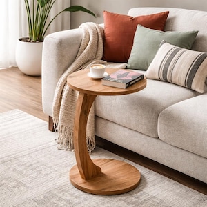 May include: A wooden side table with a circular top and base, joined by a curved support. A cup of coffee and a book are on the table. The table is beside a beige sofa with cushions and a throw.