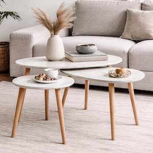 Puede incluir: Un juego de tres mesas de centro apilables con patas de madera clara y tapas blancas ovaladas y redondas. Las mesas están dispuestas frente a un sofá gris claro. Incluye un jarrón con plantas secas, libros y cuencos pequeños.