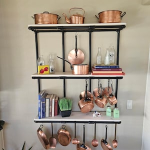 Könnte beinhalten: Ein Küchenregal mit Kupferkochgeschirr, darunter Töpfe, Pfannen und ein Kessel. Das Regal besteht aus schwarzem Metall und weißen Regalböden. Verschiedene Glasflaschen, Bücher und kleine Dekorationsgegenstände sind ebenfalls vorhanden.
