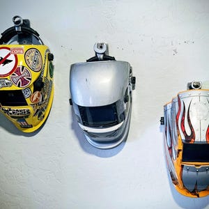 May include: Three welding helmets hang on a white wall. The helmets are yellow, silver, and orange, each with a unique design. The yellow helmet is covered in stickers, the silver one is plain, and the orange one has a flame design.