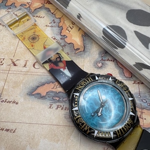 以下が含まれることがあります： 黒と金のベゼルと青い文字盤のSwatchの腕時計。時計バンドは黄色と黒で、地図と肖像画が描かれています。時計は地図の上にあります。