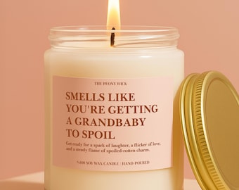 Puede incluir: Una vela en frasco de vidrio transparente con una mecha encendida y una etiqueta rosa claro. La etiqueta dice: THE PEONY WICK, SMELLS LIKE YOU'RE GETTING A GRANDBABY TO SPOIL, Get ready for a spark of laughter, a flicker of love, and a steady flame of spoiled-rotten charm. %100 SOY WAX CANDLE | HAND-POURED. Una tapa dorada se encuentra al lado del frasco.