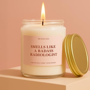 May include: A burning candle in a glass jar with a gold lid. The pink label reads "Smells Like A Badass Radiologist." The candle is hand-poured and made of soy wax.