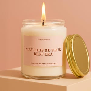 May include: A lit soy wax candle in a glass jar with a gold lid. The candle has a pink label with the text "MAY THIS BE YOUR BEST ERA" and "THE PEONY WICK". The candle is hand-poured.