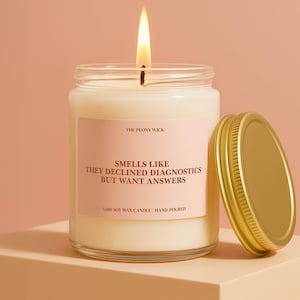 May include: A burning cream-coloured soy wax candle in a clear glass jar with a gold lid. The label reads "Smells Like They Declined Diagnostics But Want Answers." The candle is hand-poured and from The Peony Wick.