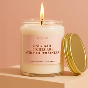 May include: A lit, white soy wax candle in a clear glass jar with a gold lid. The label reads "ONLY BAD BITCHES ARE ATHLETIC TRAINERS." The candle is hand-poured and from The Peony Wick.