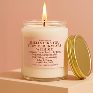 May include: A lit soy wax candle in a clear glass jar with a gold lid. The candle has a pink label with the text "Smells Like You Survived 16 Years With Me." The candle is hand-poured.